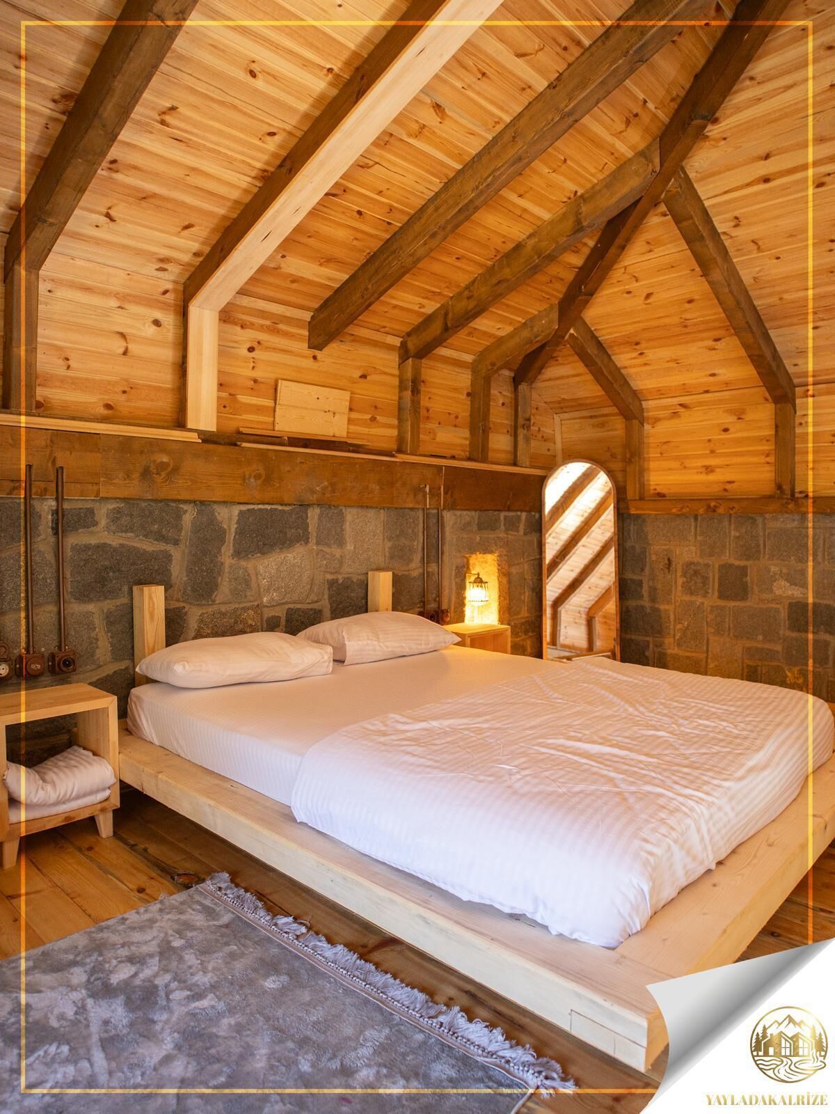 A bedroom with a stone wall, wooden ceiling beams, a bed, bedside tables, and a nightstand with a lamp.