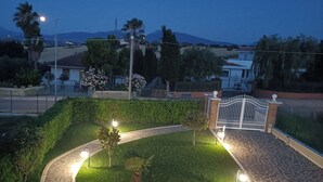 Zona de jardí paisatjista amb un camí corbat, una palmera i una entrada tancada per una reixa.