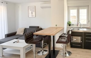 Un salon moderne comprenant un canapé, une table basse, deux tabourets de bar, une table à manger en bois et un coin cuisine avec un évier et des placards.