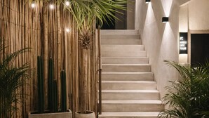 Uma escadaria leva à entrada de um prédio, ladeada por bambu e plantas em vasos, com luzes de Natal acima.