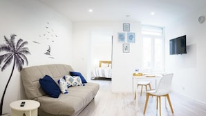 Un salon moderne avec un canapé, une petite table ronde, un fauteuil, un lit, un téléviseur et des œuvres d’art accrochées aux murs.