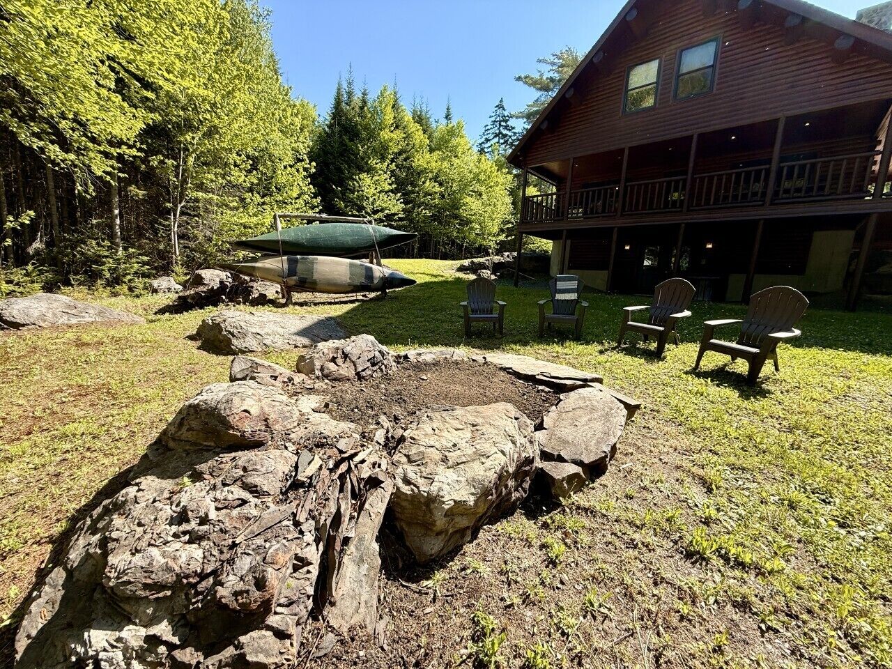 A rustic cabin with a fire pit surrounded by rocks, a deck with chairs, and a forested backdrop.