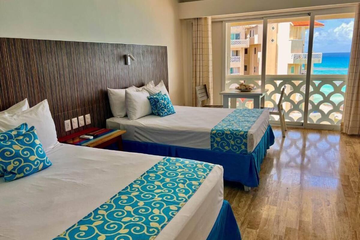 Ein Hotelzimmer mit zwei Betten, einem Holzkopfteil, einem Balkon mit Meerblick und einem kleinen Tisch mit Stuhl.
