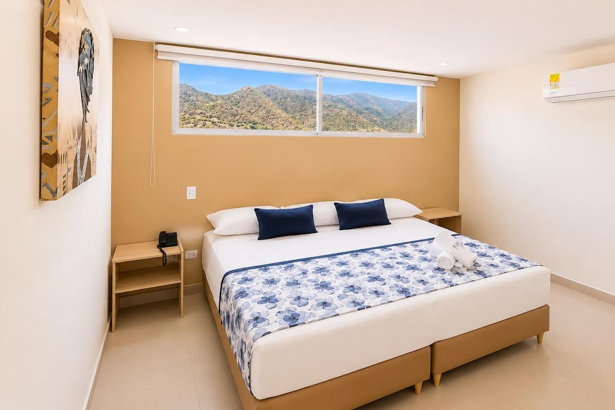 A bedroom with a bed, bedside tables, a window with a mountain view, an air conditioning unit, and a wall-mounted artwork.
