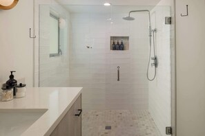 A modern bathroom with a glass-enclosed shower, a white tiled wall, and a small shelf with toiletries. - Palm Springs Movie Colony | Designer Pool Home + Private Casita, Sleeps 10 (Palm Springs)