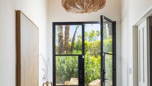 A hallway with a wooden bench, a woven light fixture, a large framed artwork, and a view through a glass door to an outdoor seating area. - Palm Springs Movie Colony | Designer Pool Home + Private Casita, Sleeps 10 (Palm Springs)