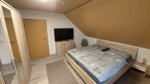 Une chambre avec un plafond en pente, un lit avec une literie à motifs, un téléviseur posé sur un meuble, une armoire en bois et une porte donnant sur une autre pièce.