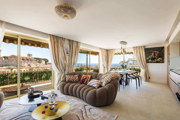 Ein modernes Wohnzimmer mit Sofa, Esstisch und Stühlen, großen Fenstern mit Vorhängen und Blick auf Gebäude und Grünflächen.