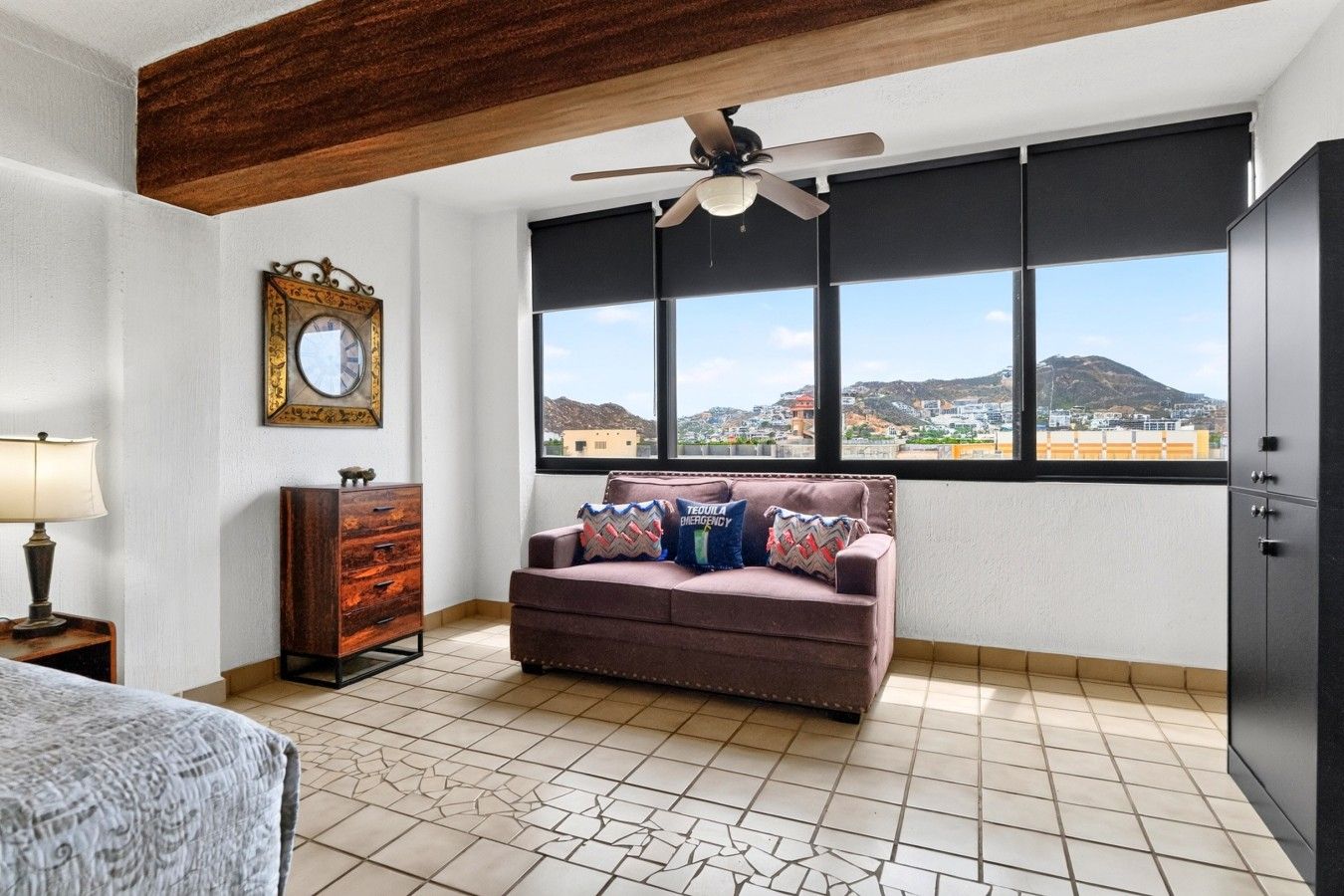A room with a sofa, a wooden dresser, a ceiling fan, and a large window with a city view.