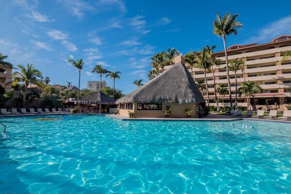Ein Resort-Pool mit einer strohgedeckten Hütte, Palmen und einem mehrstöckigen Gebäude im Hintergrund.