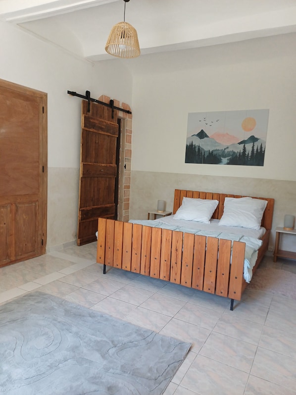 Une chambre à coucher avec un lit en bois, une grande porte en bois, un tableau accroché au mur et un luminaire suspendu.