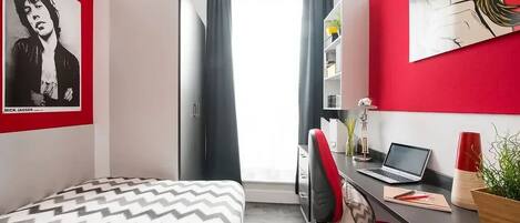 A bedroom with a bed, a desk with a laptop, a chair, and a window with curtains.