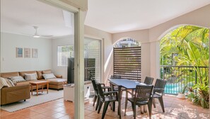 A living room with a sofa, coffee table, and TV, a dining area with a table and chairs, and a balcony with a view of a pool and tropical plants. - Sanctuary Lake Apartment 54 - Peaceful, Beachside Currumbin Oasis - Min. 3 Night Stays! (Currumbin)