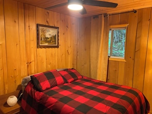 A wooden cabin room with a bed, a painting, a window, and a ceiling fan.