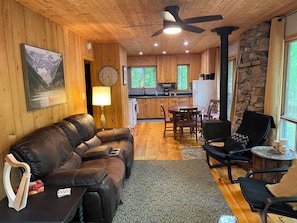 A cozy cabin interior with a leather sofa, dining table, chairs, a kitchen area, and a stone wall.