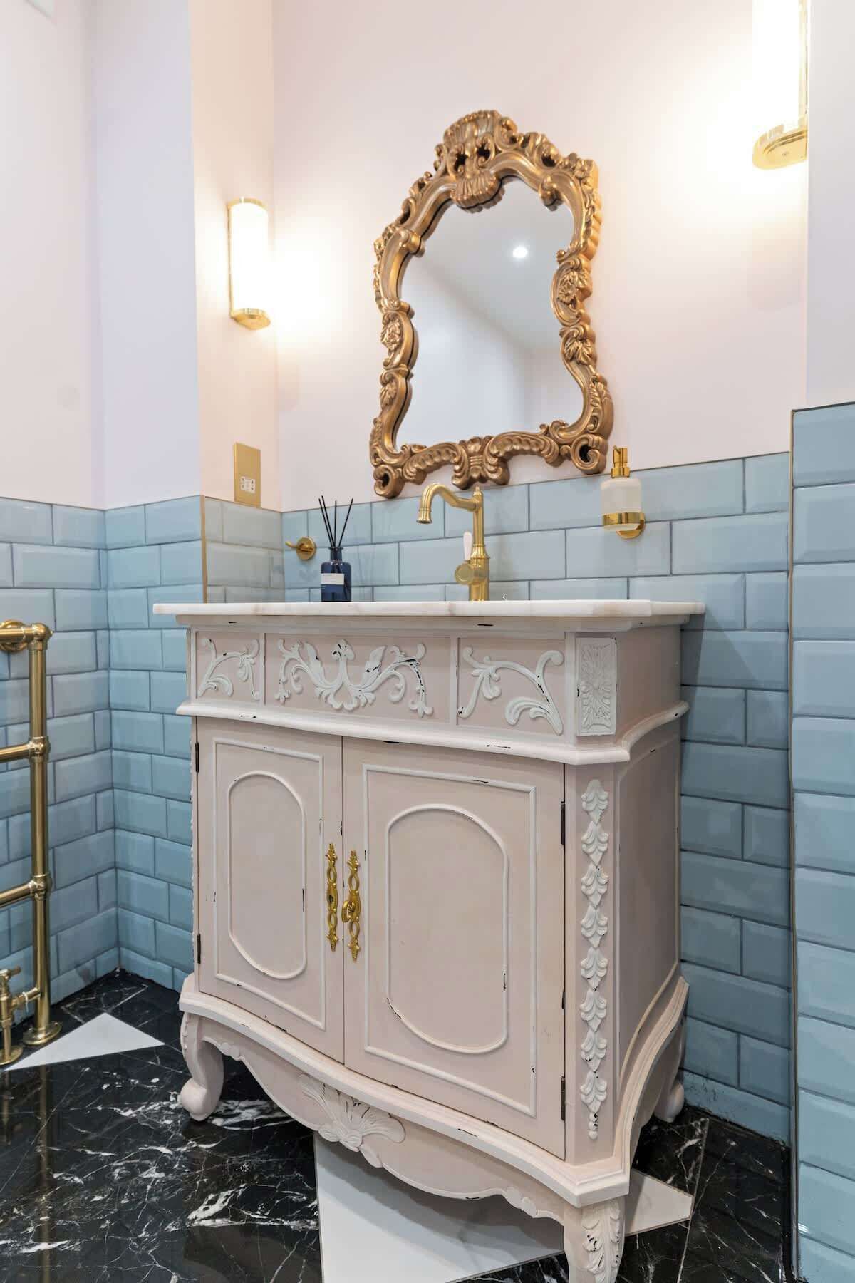 Un baño con tocador blanco, espejo dorado y paredes con azulejos azules.