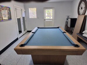 A pool table in a room with a door, windows, and a clock on the wall.