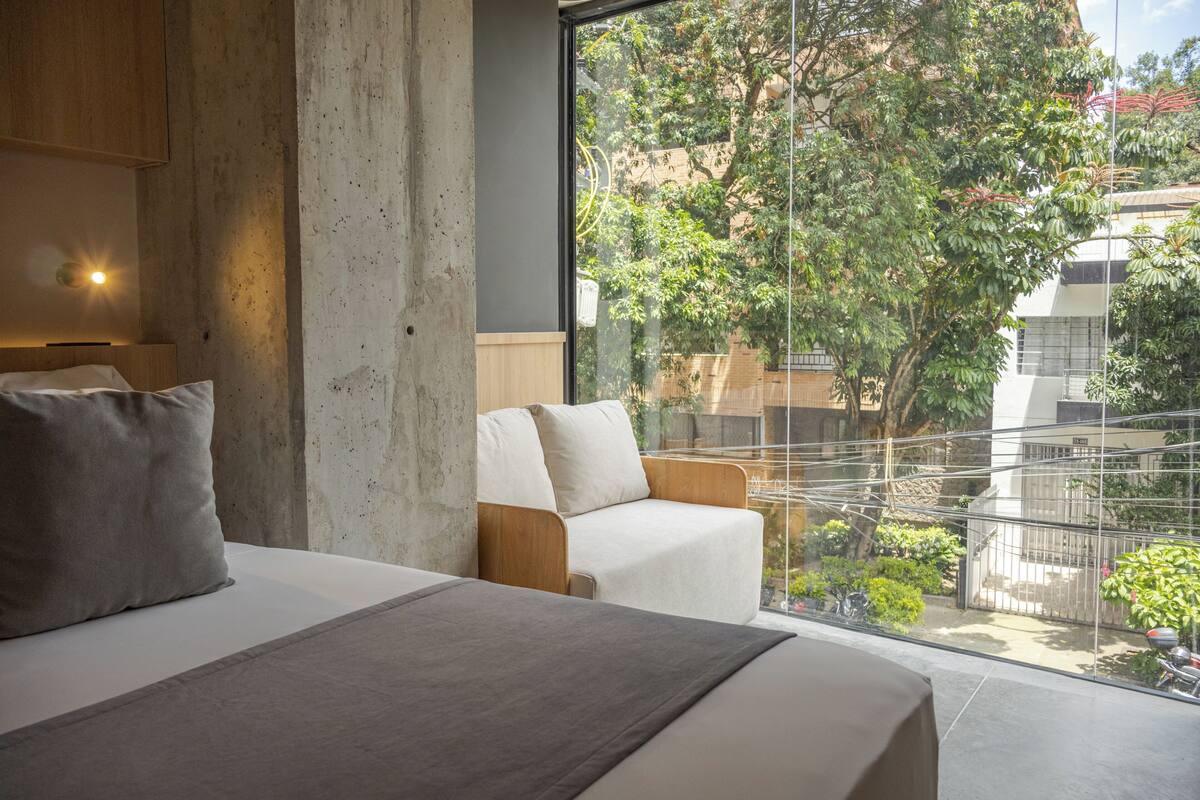 Un dormitorio moderno con una cama grande, un cabecero de madera, un sofá blanco y un balcón con vistas a zonas verdes y edificios.