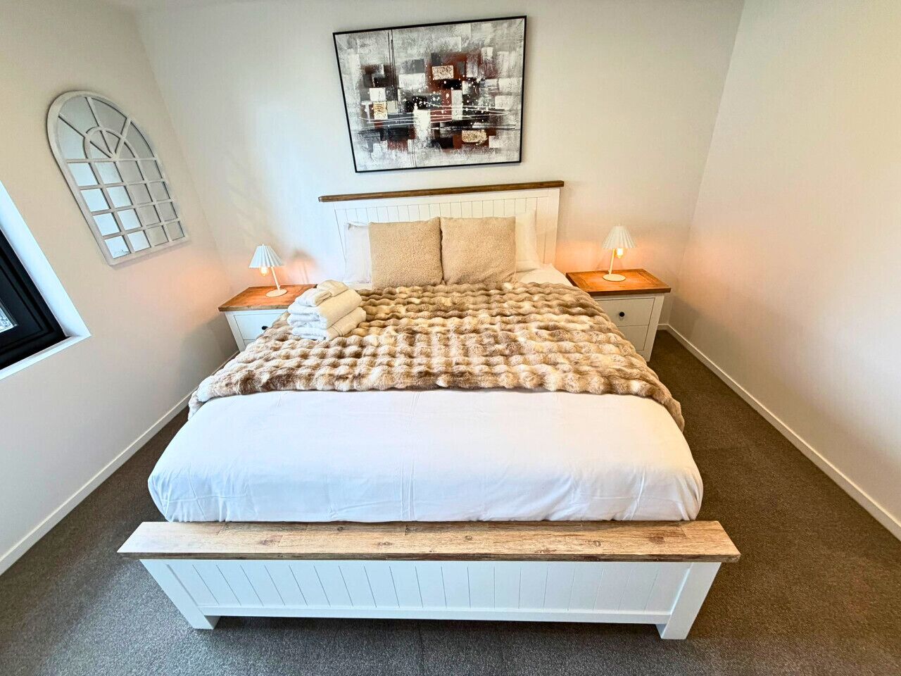 A bedroom with a bed, two bedside tables, a headboard, a window, and a wall-mounted artwork.