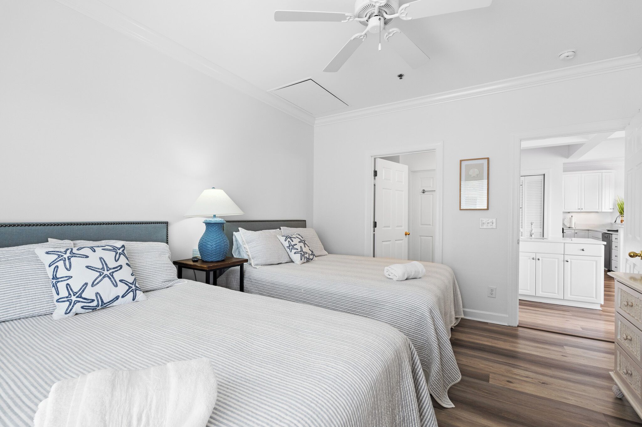 A hotel room with two beds, a ceiling fan, a nightstand with a lamp, and a view into the kitchen area.