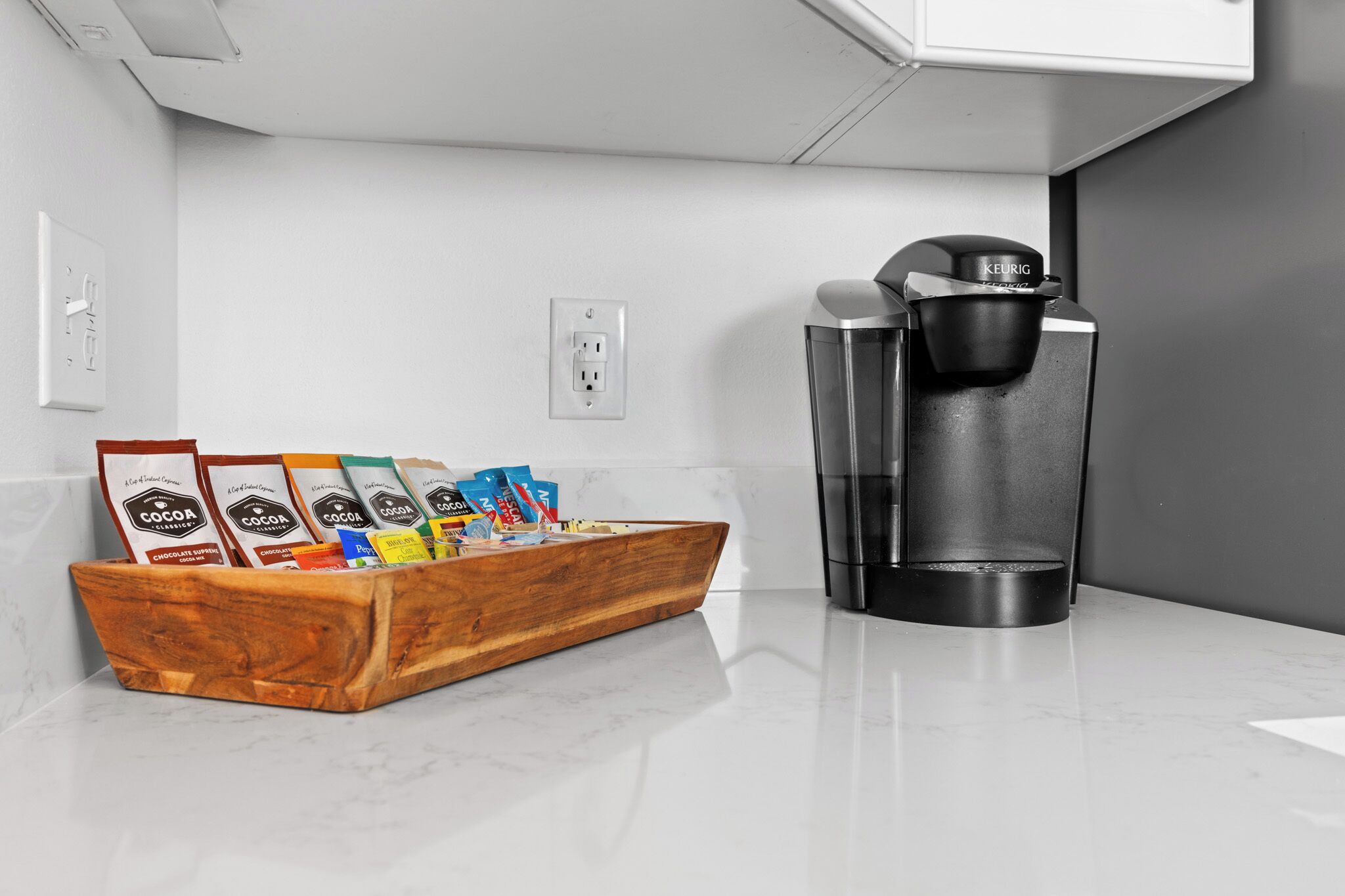 A kitchen with a coffee maker, a wooden tray with coffee pods, and a white countertop.