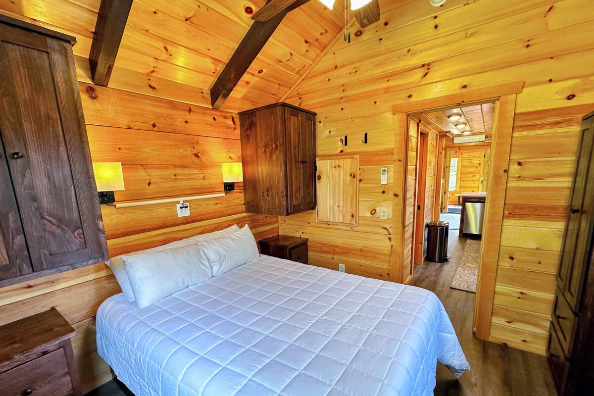 Une chambre à coucher dans une cabane en bois, avec un lit, une table de chevet, une armoire et une salle de bain visible à travers une porte ouverte.