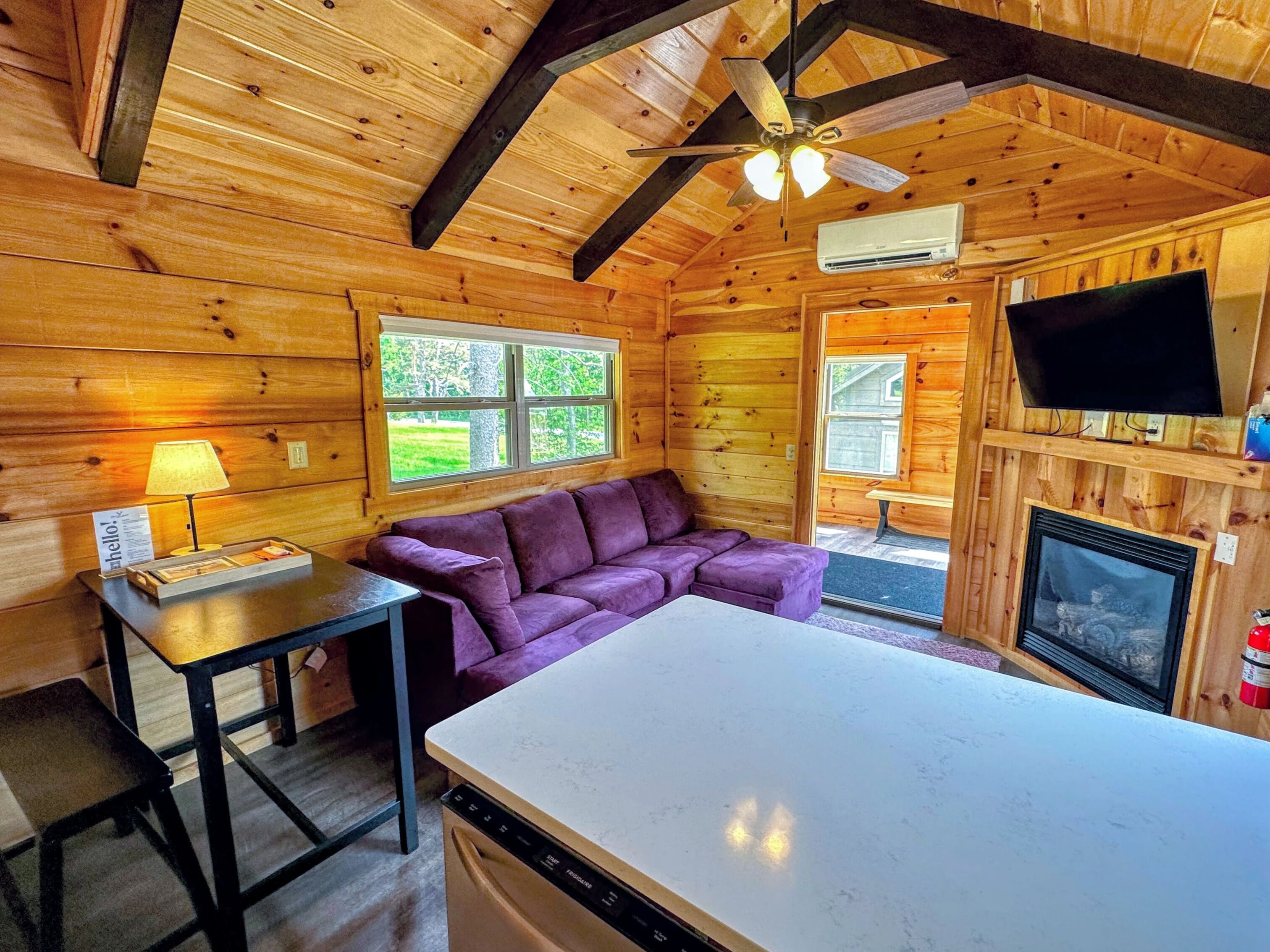 A wooden cabin interior with a sofa, a table, a ceiling fan, a fireplace, and a television.
