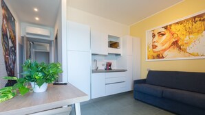 A modern kitchen with white cabinets, a dining table with a potted plant, a sofa, and a large abstract painting on the wall.