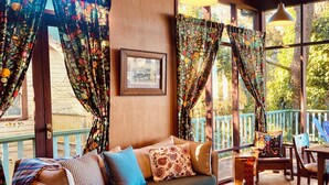 Ein gemütliches Zimmer mit einer Couch, einem Tisch und einem Sessel. Ein Fenster mit Vorhängen und ein Bilderrahmen sind an der Wand zu sehen.