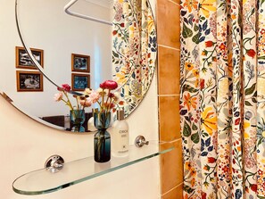 Une salle de bain avec une étagère en verre, un miroir, des rideaux à motifs floraux et des tableaux encadrés accrochés au mur.