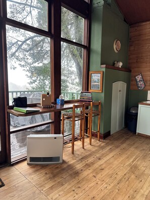 Een kamer met een groot raam, houten vloer, een tafel met boeken, een hangklok en een lijst met een afbeelding.
