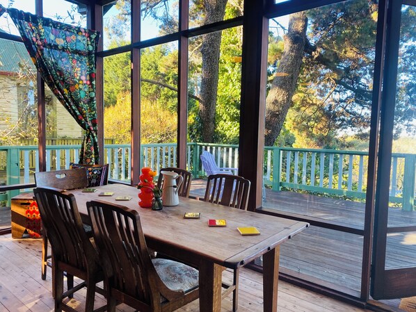 A wooden deck with a table and chairs, surrounded by glass walls, overlooking a green deck and trees. - Cosy Blue Mountains Cottage | Fireplace • Walk to Town • Relax & Unwind (Katoomba)