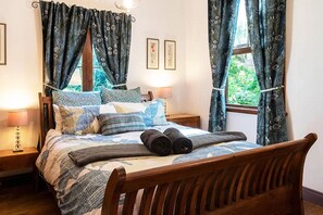 Ein Schlafzimmer mit einem hölzernen Bett, mehreren Kissen, einem zusammengerollten Handtuch, zwei Nachttischlampen und Vorhängen.