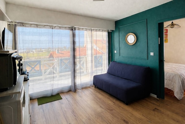 A room with a sofa, a bed, a window with curtains, a mirror, and a view of buildings outside. - Cozy one-bedroom apartment for 2  just steps from the ocean (MIMIZAN PLAGE)