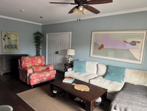 Ein Wohnzimmer mit Deckenventilator, einem roten, mit Blumenmuster versehenen Sessel, einem weißen Sofa mit blauen Kissen, einem Holztisch und einem gerahmten abstrakten Gemälde an der Wand.