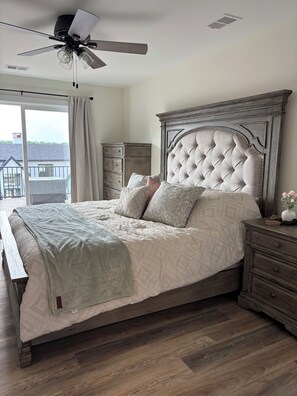 A bedroom with a large bed, a ceiling fan, a dresser, and a view of the outside.