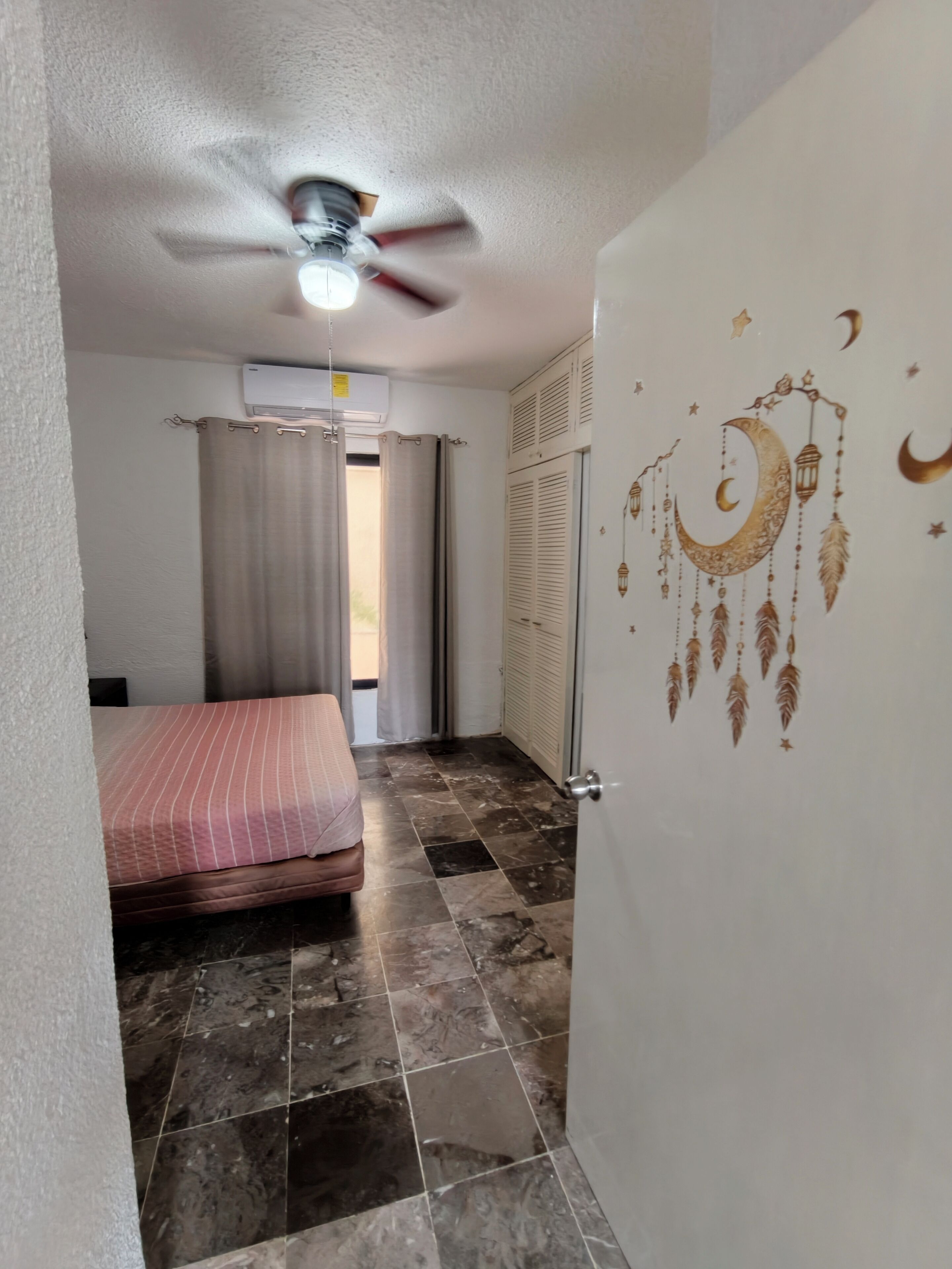 A bedroom with a bed, a ceiling fan, a window with curtains, and a wall with a decorative design.