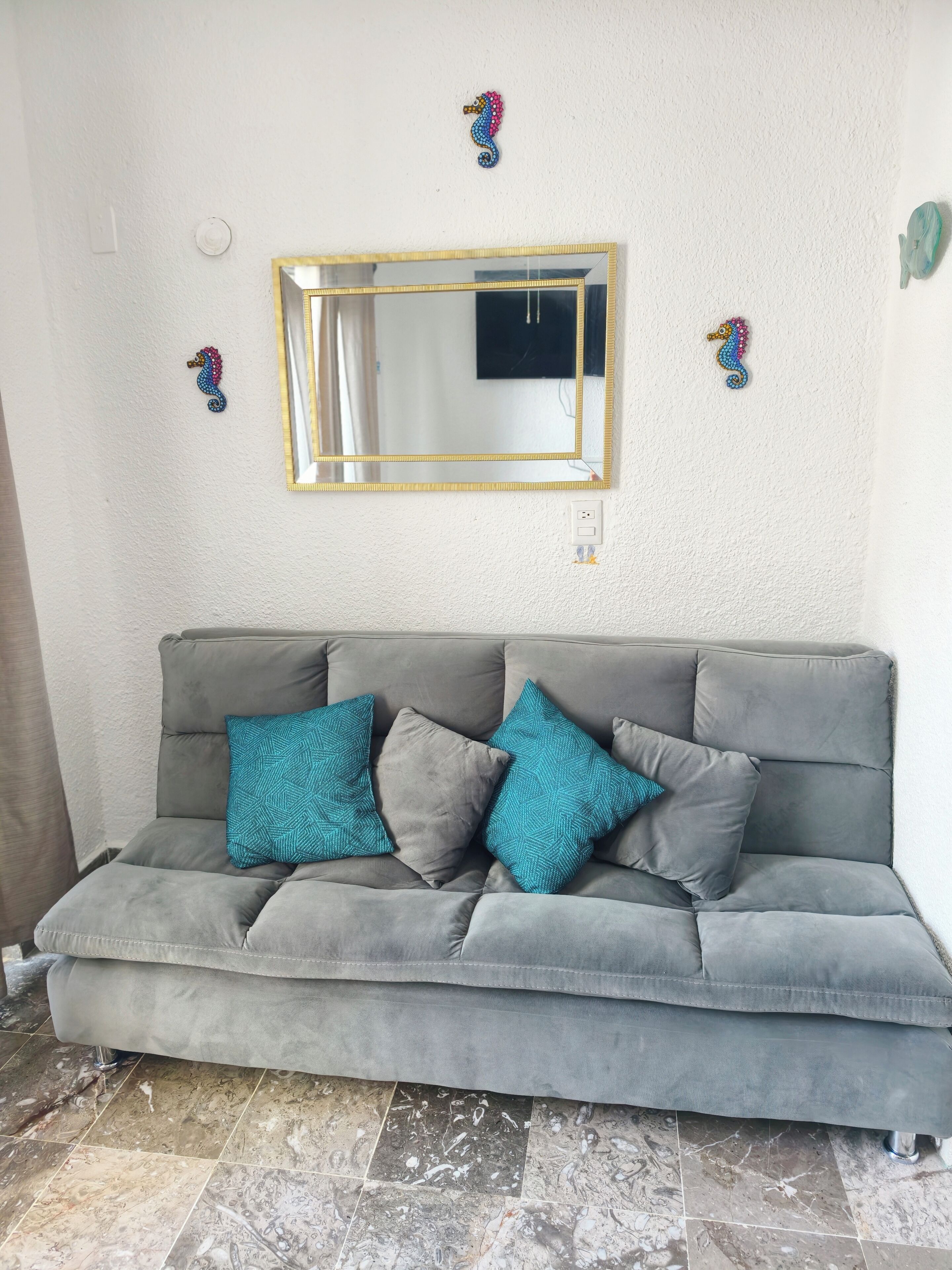 Un sofá gris con cojines de estampado azul verdoso, un espejo con marco dorado y decoración de caballitos de mar en la pared.