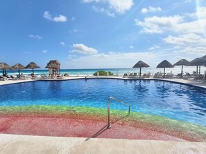A pool with a clear blue water surface, surrounded by lounge chairs and thatched umbrellas, with a view of the ocean. -  Cancún Caribe Soul studio /walk on beach/ Top Location (Cancún)