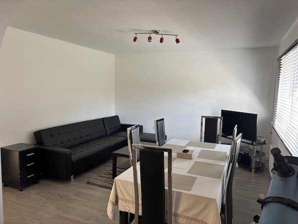 Ein modernes Wohnzimmer mit einer schwarzen Couch, einem Esstisch mit Stühlen, einem Fernseher und einer Deckenleuchte.