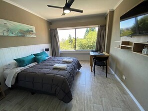 A bedroom with a bed, a desk, a chair, a ceiling fan, a TV, and a window with curtains.
