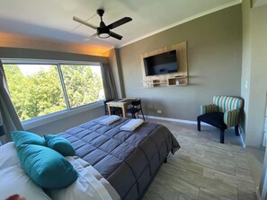 A bedroom with a bed, a desk, a chair, a TV, and a view of trees.