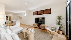 A modern living room with a sofa, coffee table, TV, and a dining area with a kitchen in the background. - Unit 1, 9 Hill Street, Sunshine Beach (Sunshine Beach)