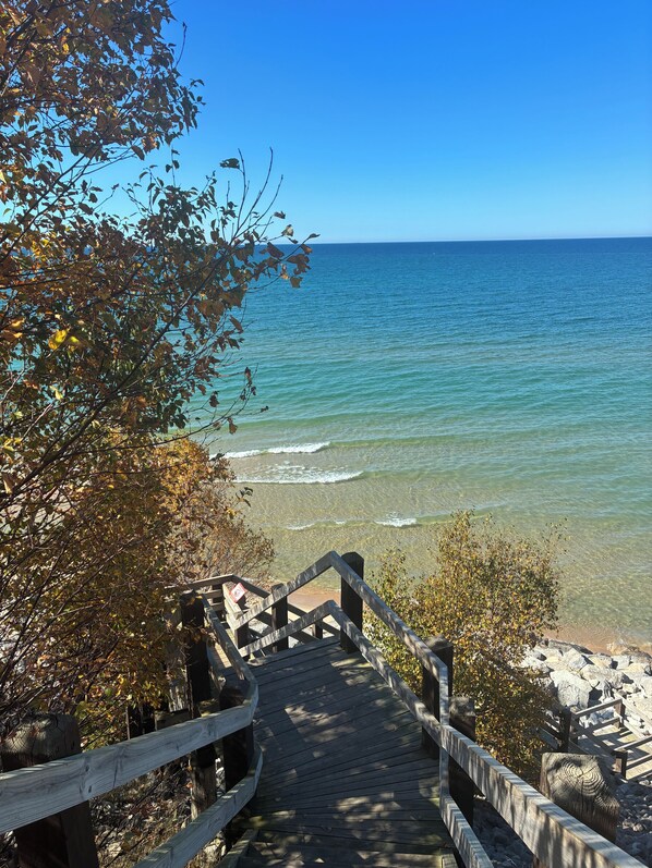 A wooden staircase leading to a beach with clear blue water and a clear sky. - Manistee Harbor Retreat · Lake Michigan Access · Indoor Pool · Sleeps 8 (Manistee)