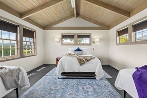 A bedroom with a sloped ceiling, a bed with a white comforter and a blue pillow, two windows with blinds, and a view of greenery outside. - Yakima Valley Views with Pool and Sauna (Yakima)