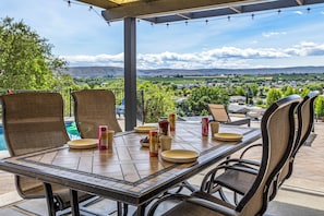 A covered patio with a table set for dining, surrounded by chairs, and a scenic view of a valley. - Yakima Valley Views with Pool and Sauna (Yakima)