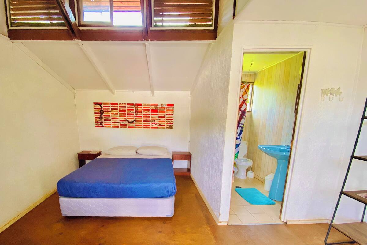 A small, compact room with a bed, a nightstand, a wall-mounted artwork, a bathroom with a sink, and a door leading to another room.