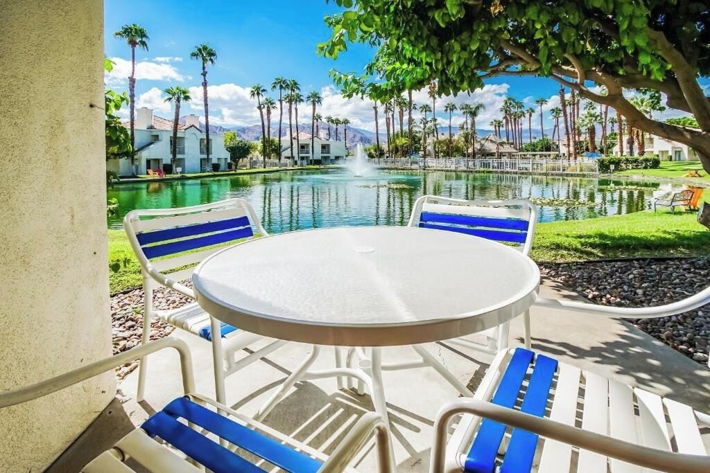 Un espace aménagé au bord de la piscine, avec une table ronde et des chaises, une fontaine, et une vue sur les palmiers et les bâtiments.