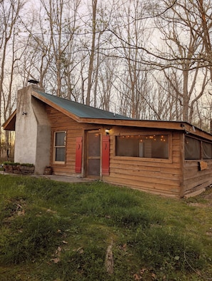 Eine rustikale Holzhütte mit geneigtem Dach, Schornstein und einer kleinen Veranda mit roten Fensterläden.