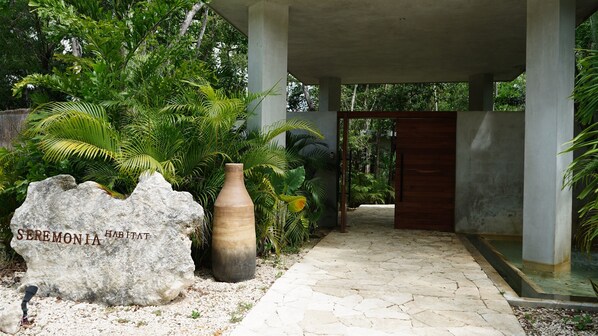 A stone pathway leading to a wooden door, surrounded by lush greenery and a large rock with 'SEREMONIA HABITAT' engraved. - Villa · Pools Privadas · Rooftop · Selva Tulum (77760)